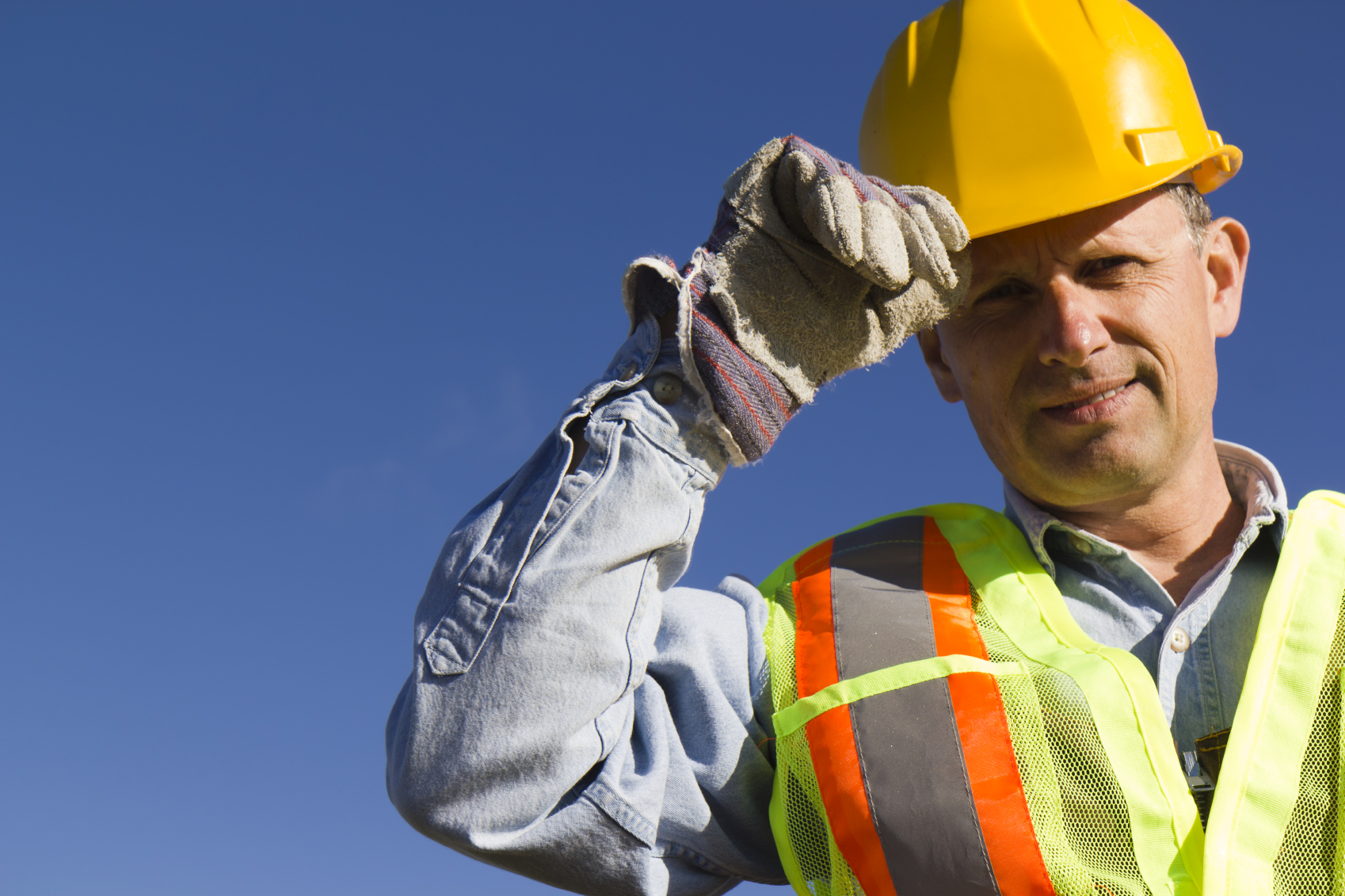 A construction worker wearing a yellow hard hat and high-visibility vest, tipping his hat under a clear blue sky.
