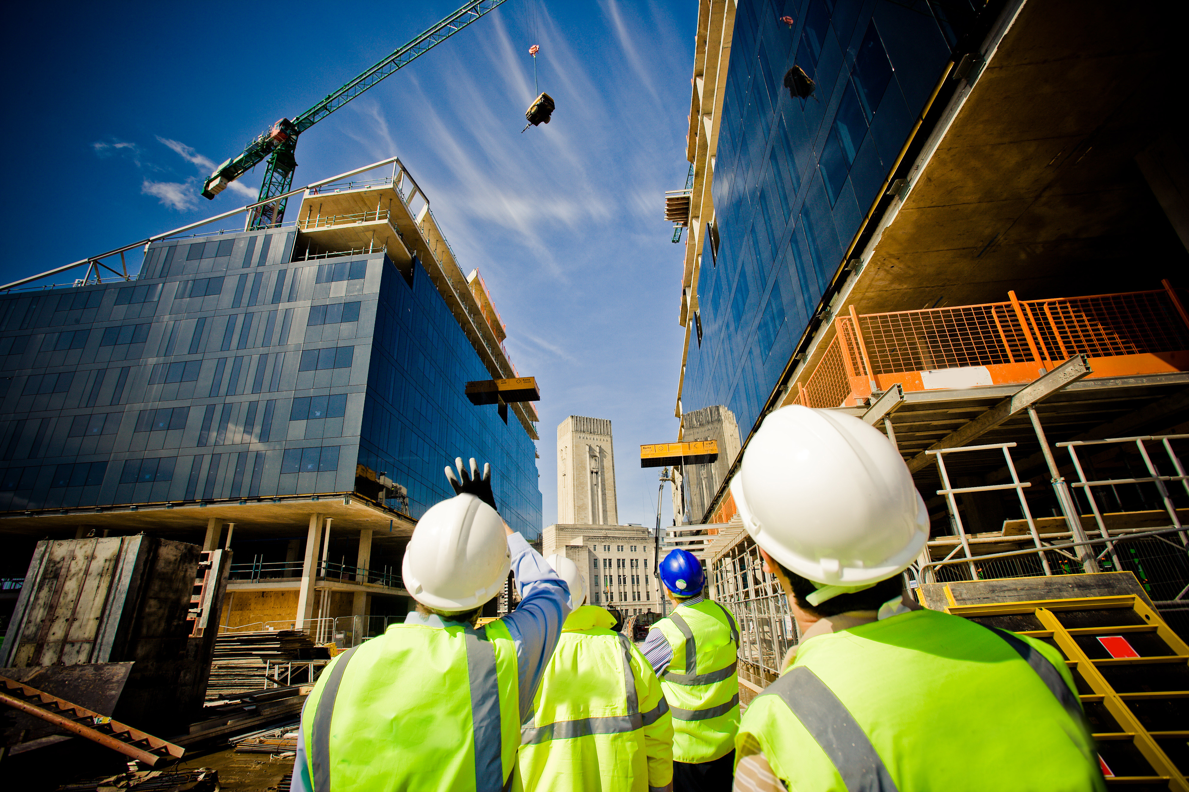 A group of construction workers in high-visibility vests and helmets on a construction site, observing a crane lifting materials.