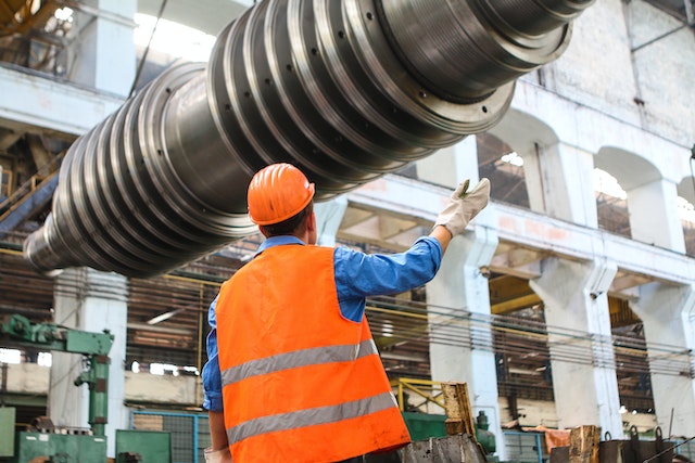 A factory worker wearing an orange safety vest and hard hat, directing the movement of a large industrial machine part.